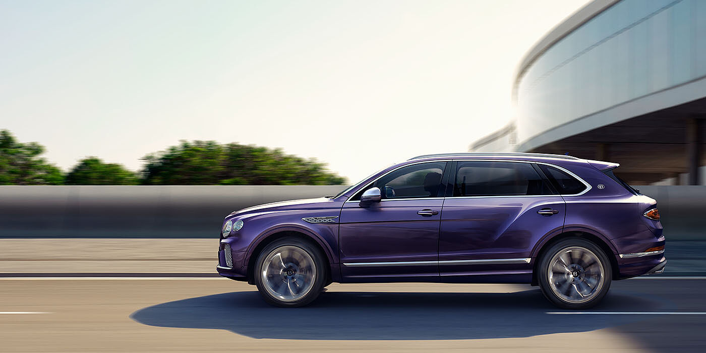 Bentley Surrey Bentley Bentayga Extended Wheelbase Mulliner luxury SUV profile in Tanzanite Purple paint, driving on an country road past trees.