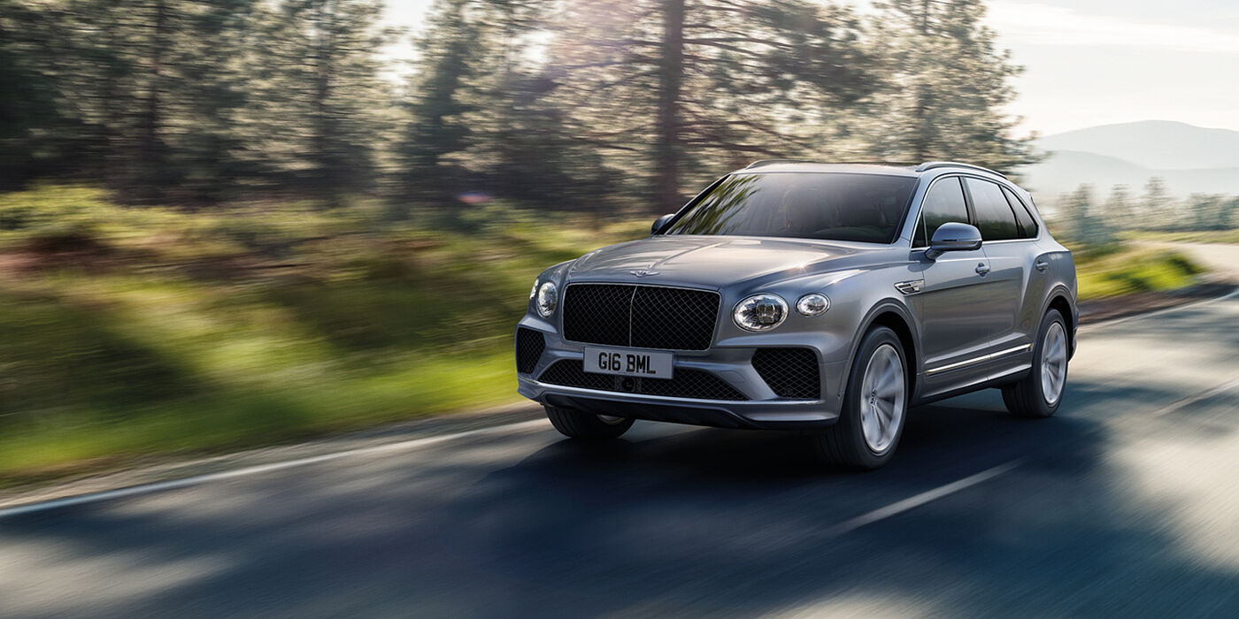 Bentley Surrey Bentley Bentayga luxury SUV front three quarter in Moonbeam paint, driving on an country road past trees.
