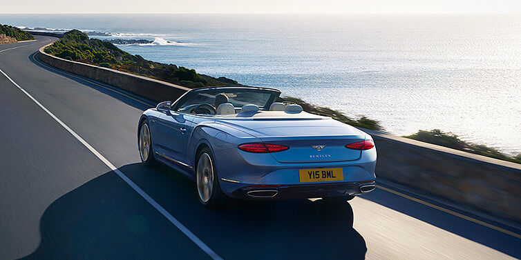 Bentley Surrey New Bentley Continental GTC Azure convertible rear view in Light Windsor blue paint with 22 inch ten spoke wheels driving dynamically on an ocean road in the sunshine