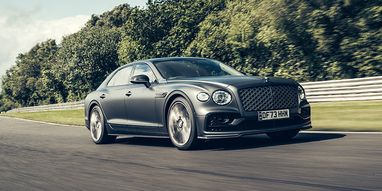 Bentley Surrey Bentley Flying Spur Mulliner sedan front three quarter in dark grey paint driving dynamically by some trees on a track