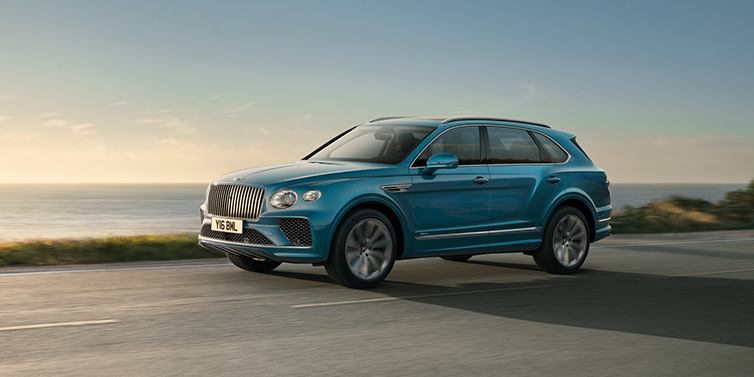 Bentley Surrey Bentley Bentayga Azure SUV in Topaz blue paint driving dynamically by the ocean with 22 inch 10 spoke directional wheels