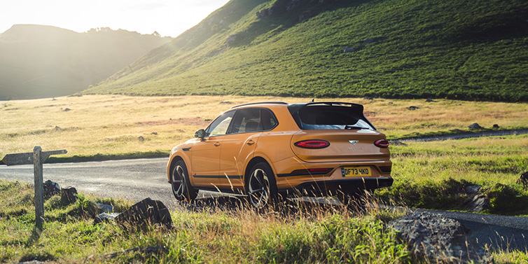 Bentley Surrey Bentley Bentayga S SUV rear three quarter in Sunburst Gold paint on a road surrounded by green hills