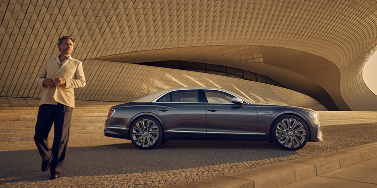 Bentley Surrey Bentley Flying Spur Mulliner sedan in Rose Gold over Magnetic duotone paint parked by a curved wall with person in suit jacket standing on the left